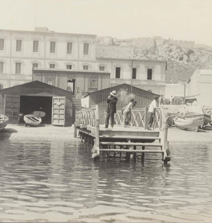 Primitiva estación marítima de la Junta de Obras del Puerto en una imagen fechada en 1909 realizada por el fotógrafo Antonio Mateos. En primer plano se ve el embarcadero, las barcas y las casetas, muy cercada del proyecto del Balneario Neptuno. Al fondo, la Alcazaba.