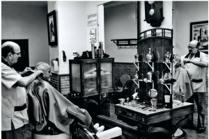 Imágenes de la barbería Mora con José Mora atendiendo a un cliente (arriba) y el estado actual, deshabitado por cierre, de la barbería (abajo).