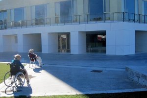 Una mujer en silla de ruedas toma el sol junto a un hombre a las puertas de una residencia de mayores.