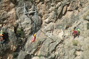 Una persona se tira en tirolina en una montaña mientras otras siguen una via ferrata por la ladera de otra.