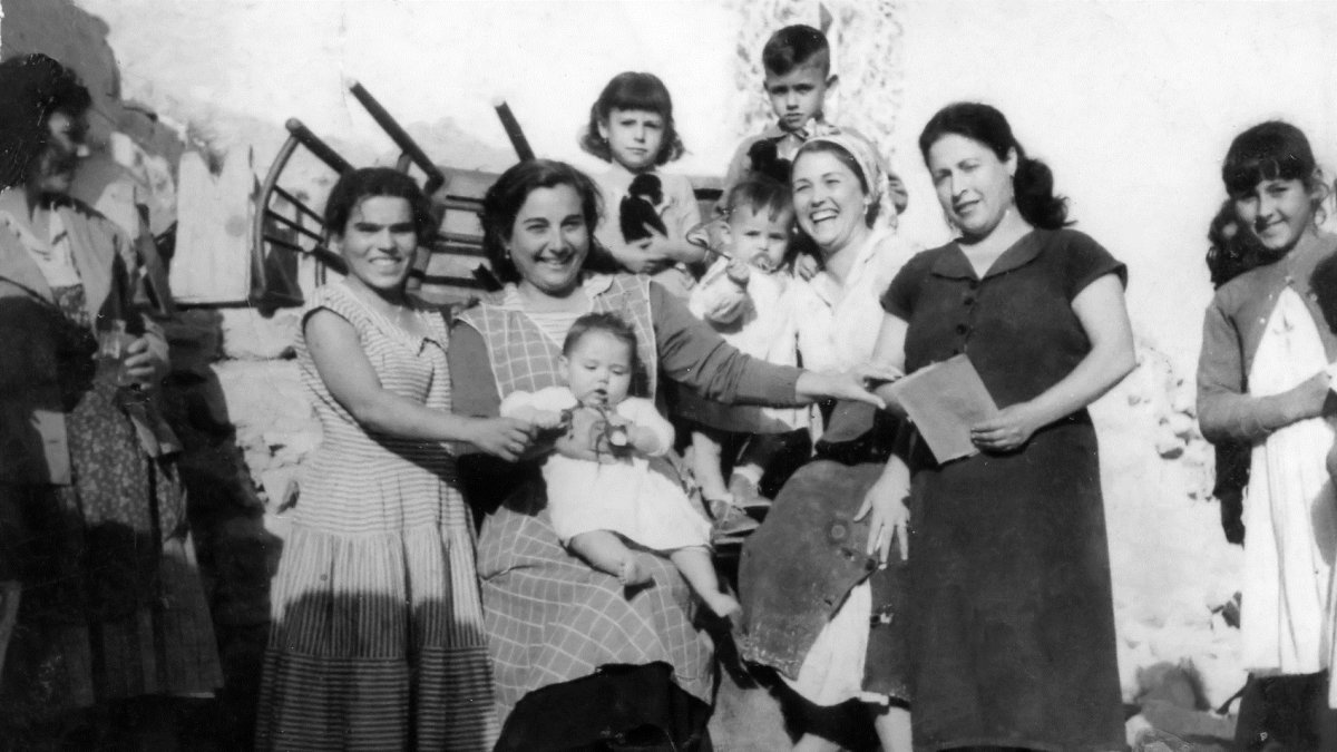 La señora matilde  con vestido negro y una libreta en la mano, en la puerta de la tienda, donde los vecinos y los clientes se solían sentar aprovecha