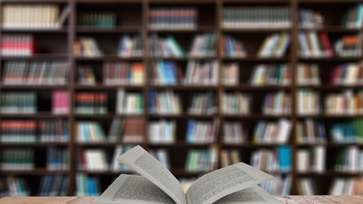 Libro abierto con una estantería llena de ejemplares apilados al fondo.