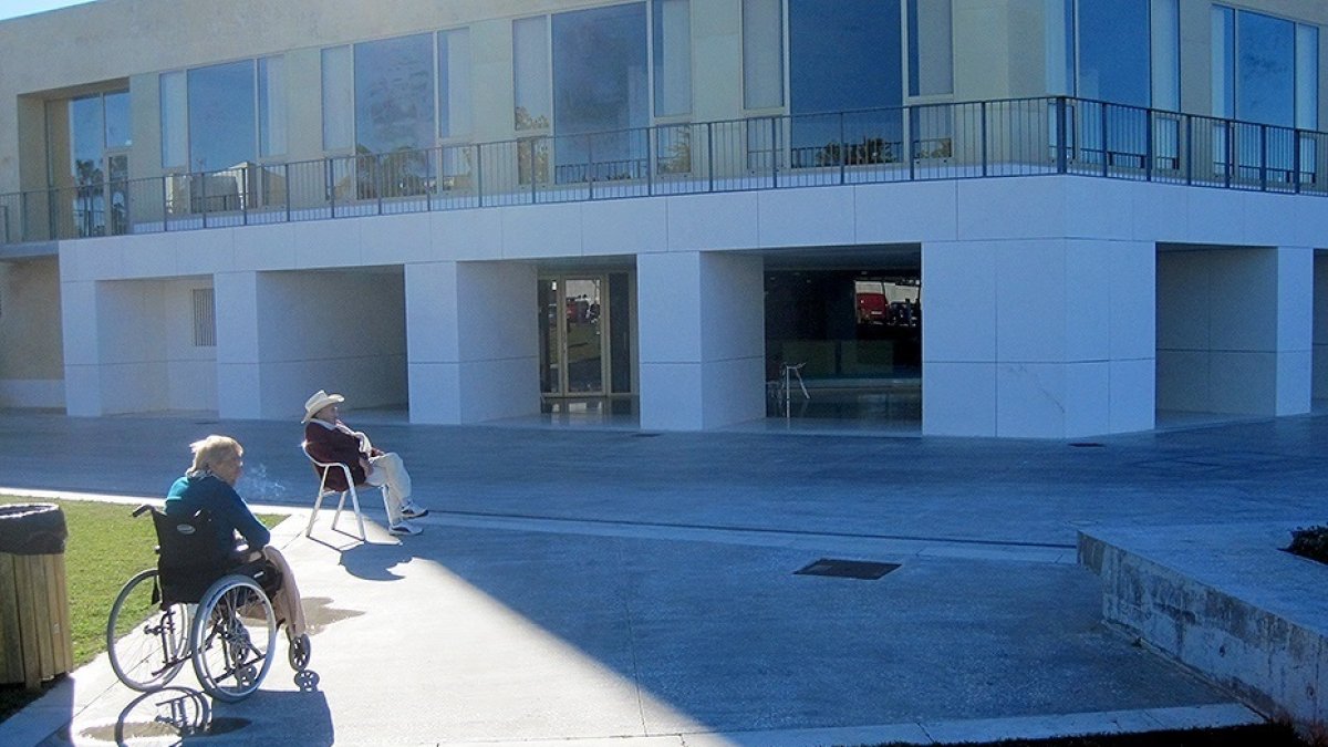 Una mujer en silla de ruedas toma el sol junto a un hombre a las puertas de una residencia de mayores.