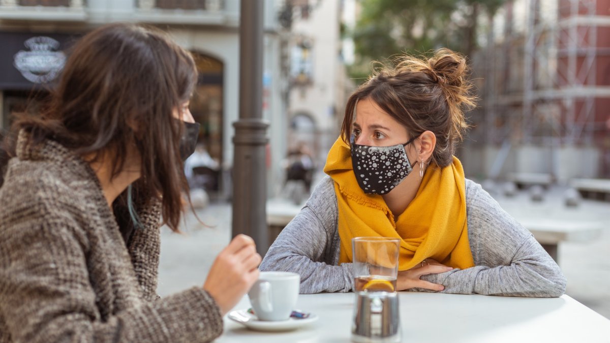 Dos mujeres toman café con mascarilla en una terraza.